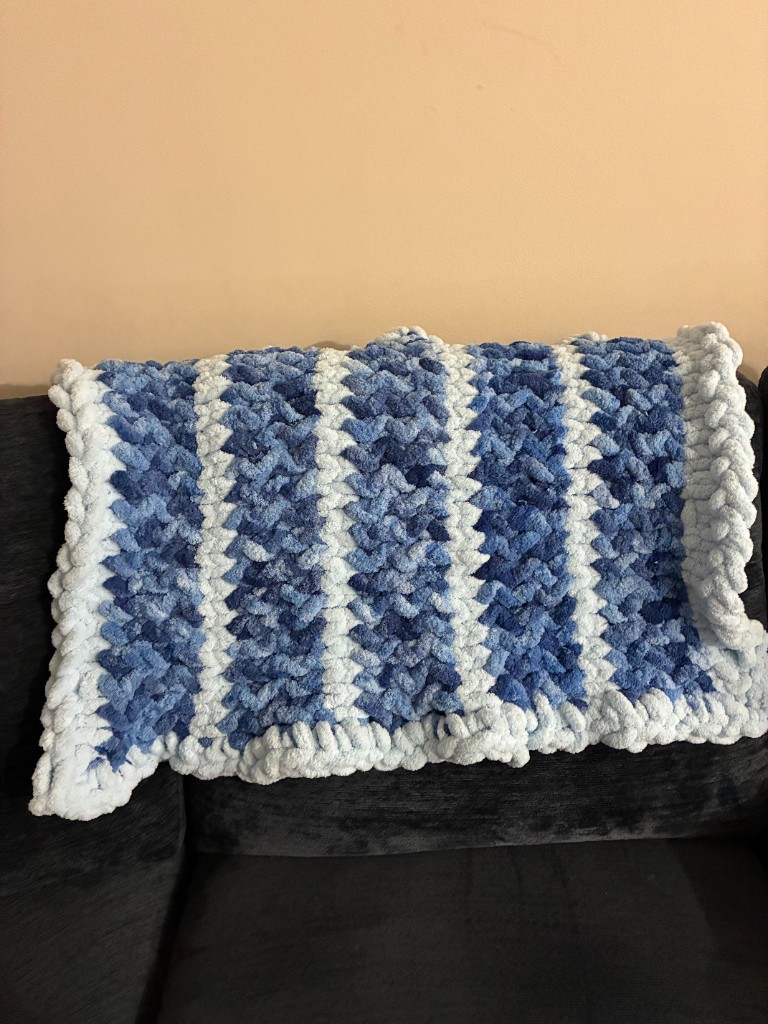 A chunky finger-knit blanket in blue and white stripes, draped over a dark sofa.