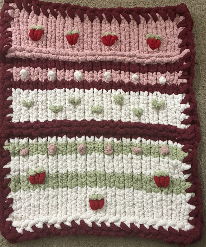 A chunky finger-knit blanket with pink, cream, sage, and berry stripes and knitted strawberries.