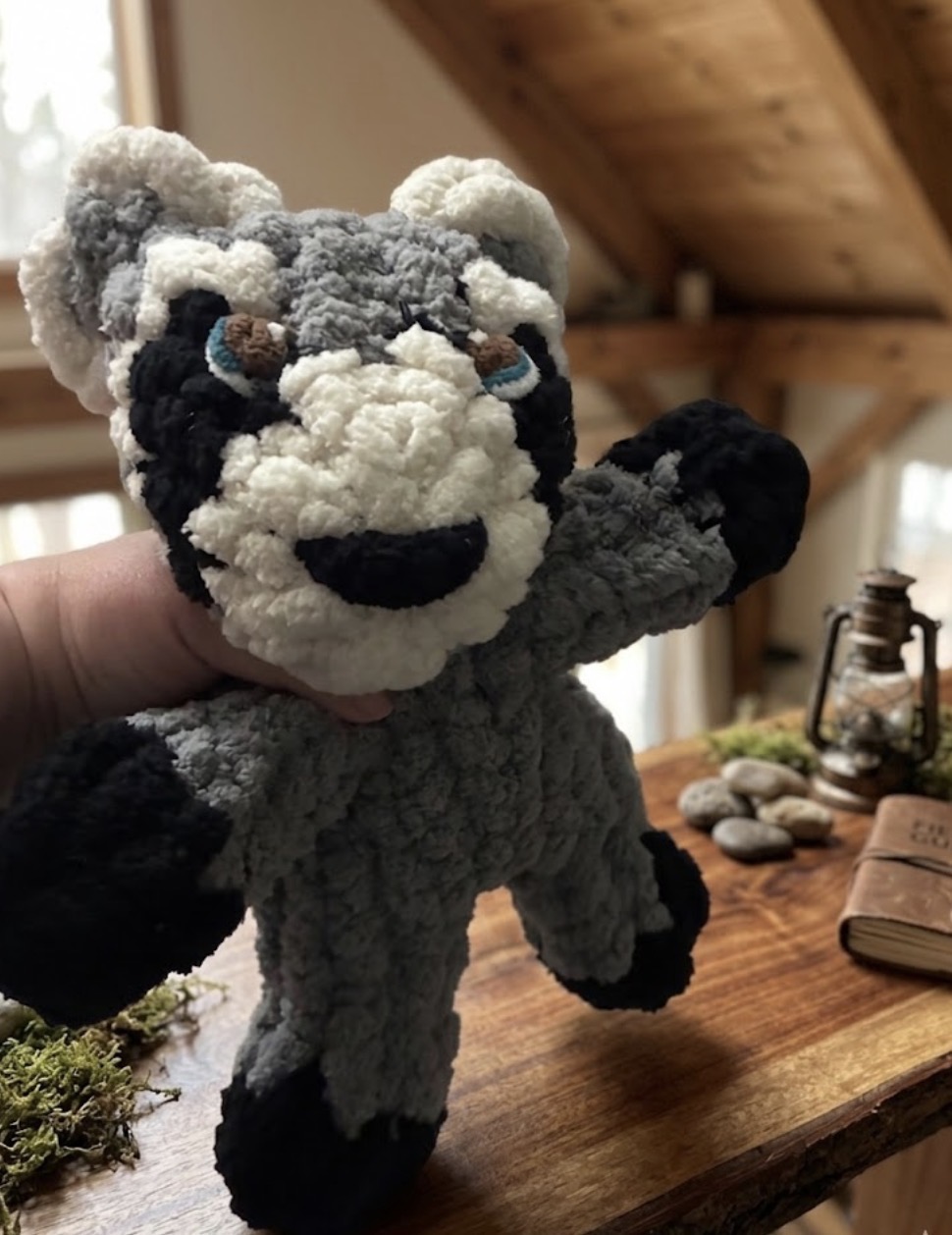 A gray and white finger-knit raccoon stuffie held in a wood-beamed studio.