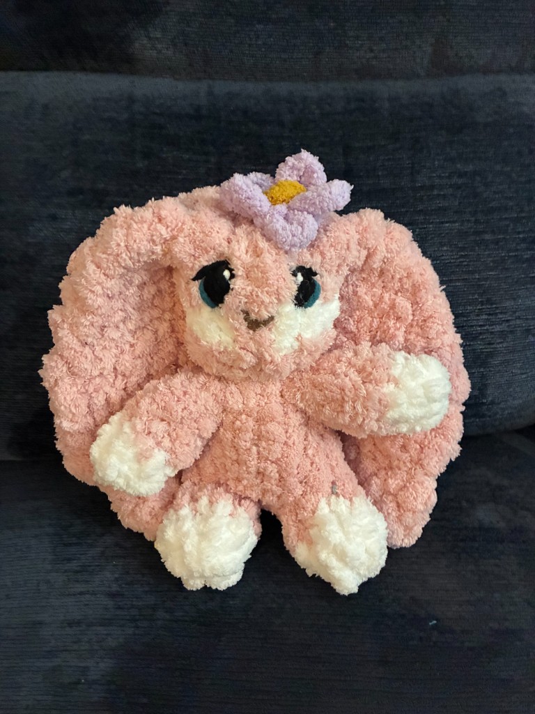 A pink finger-knit bunny stuffie with a lavender flower, sitting on a dark sofa.