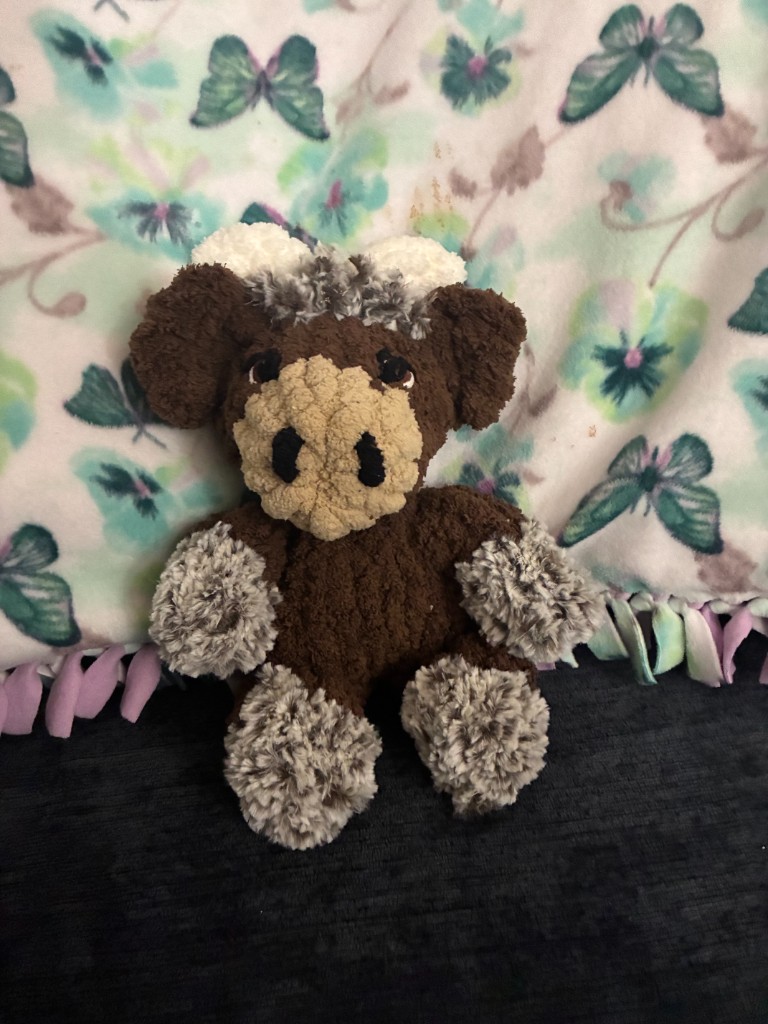A brown finger-knit cow stuffie with fuzzy gray hooves, sitting against a floral blanket.